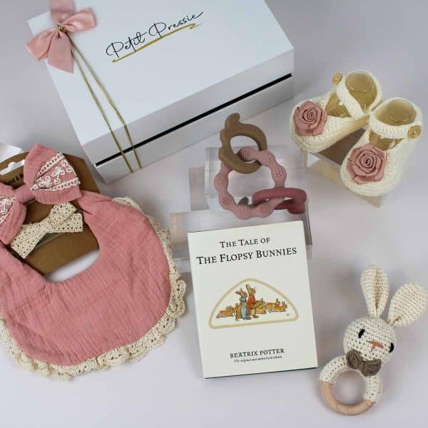 Baby girl gift box flat lay with Petit Pressie branding, pink bib, bows, book, teether, shoes, and bunny rattle