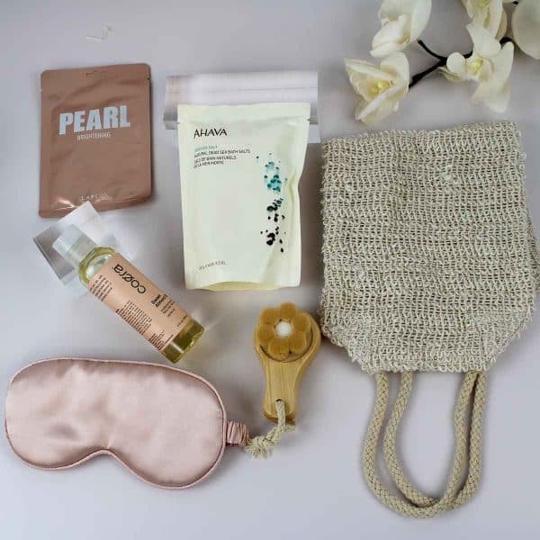 Close-up of a spa gift box featuring bath salts, almond oil, a brightening face mask, a facial brush, a pink satin eye mask, and an exfoliating mitt, perfect for gifting.