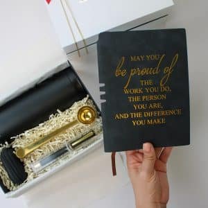 Motivational gift box with black tumbler, gold coffee scoop, pen, carrying case, and a black journal that reads “May you be proud of the work you do, the person you are, and the difference you make”