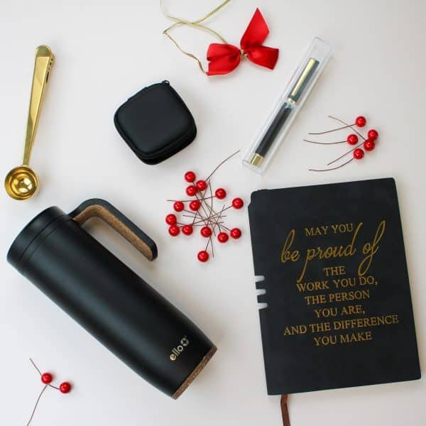 Holiday flat lay with motivational journal, black Ello tumbler, gold coffee scoop, pen, red berries, and gift ribbon on white background