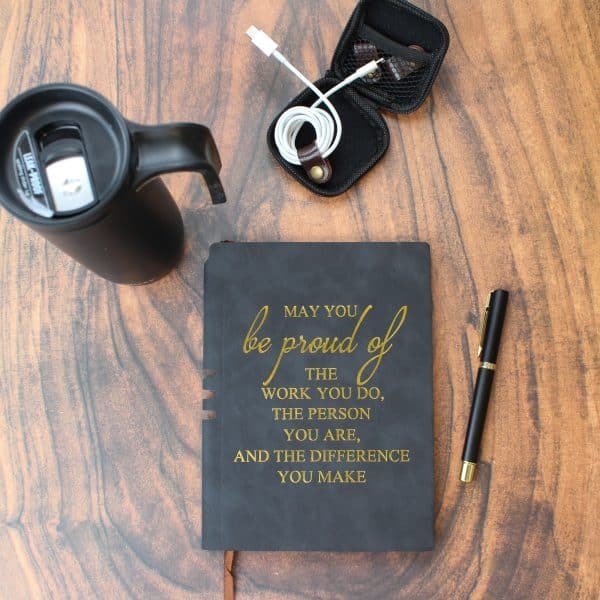 Motivational flat lay with black journal featuring gold quote, black tumbler, pen, and cable case on wooden desk