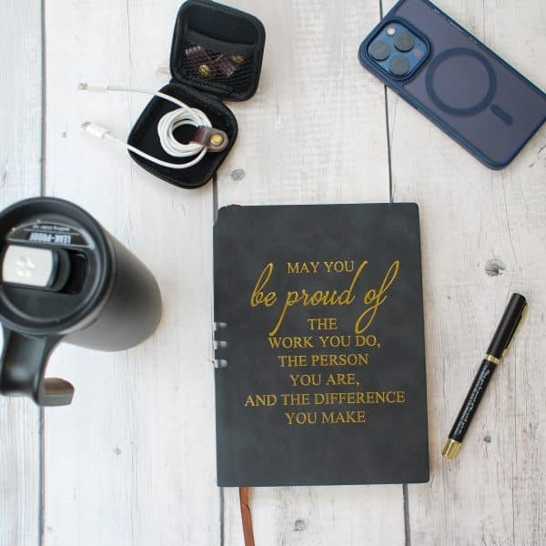 Flat lay of motivational journal with gold quote, black tumbler, earbuds case, charging cable, smartphone, and black pen on white wooden surface