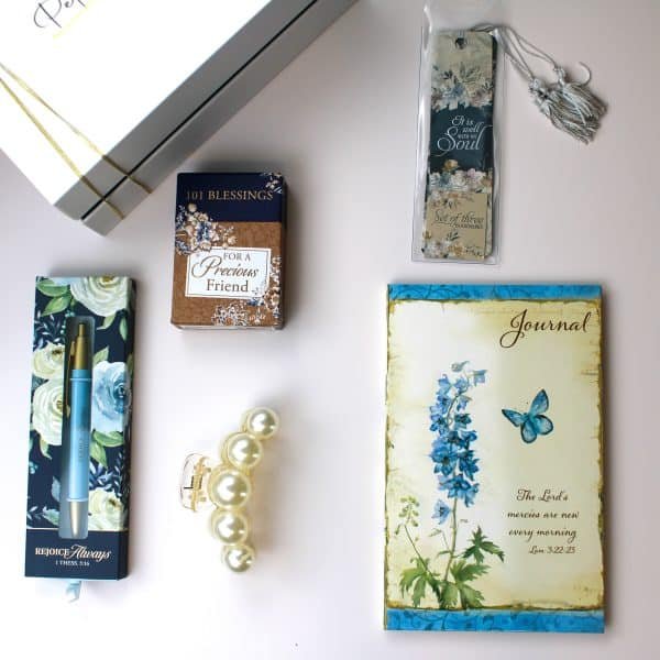 Christian faith-inspired gift flat lay with floral journal, scripture bookmark set, pen with Bible verse, 101 blessings card deck, pearl hair clip, and Petit Pressie box