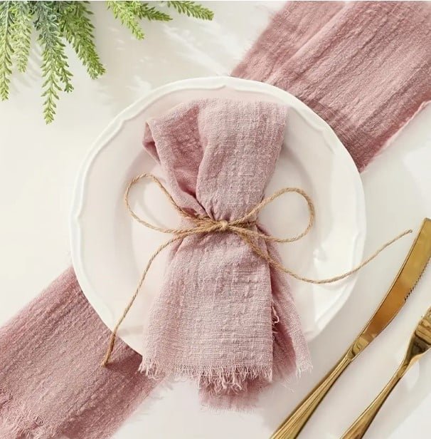 Rustic table setting with soft mauve gauze napkin tied with twine on white ceramic plate, mauve table runner, and gold cutlery