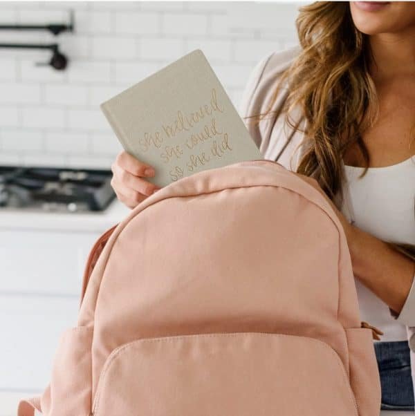 Woman placing a beige journal with gold text that reads “She believed she could so she did” into a soft pink backpack.