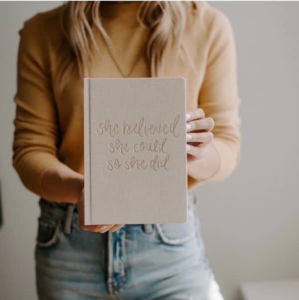 Woman in mustard sweater and jeans holding a beige journal with gold lettering that reads “She believed she could so she did.”