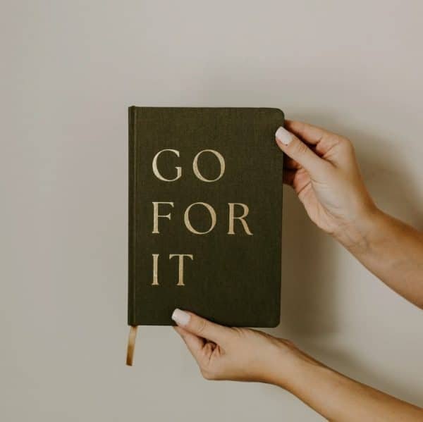 Hands holding a dark olive green journal with gold embossed text that reads “GO FOR IT” against a neutral background.
