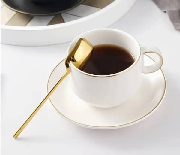 White porcelain coffee cup with gold rim and saucer, served with a modern gold spoon resting on the edge
