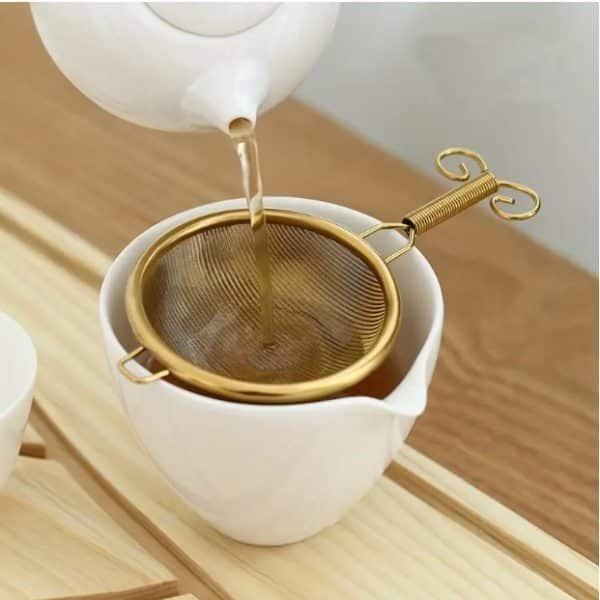 Close-up of tea being poured from white teapot through elegant gold mesh strainer into white ceramic cup on wooden tray