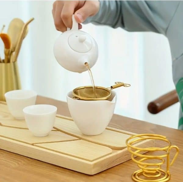 Pouring tea from white ceramic teapot through gold mesh strainer into white teacup on wooden tray with matching accessories
