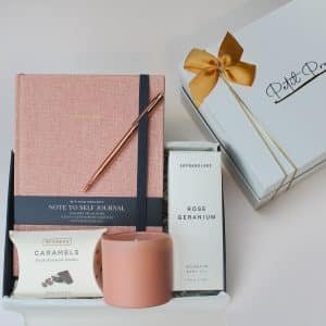 Faith-based gift box with journal, rose gold pen, rose geranium body oil, mocha caramels, and pink candle inside a white gift box with gold bow