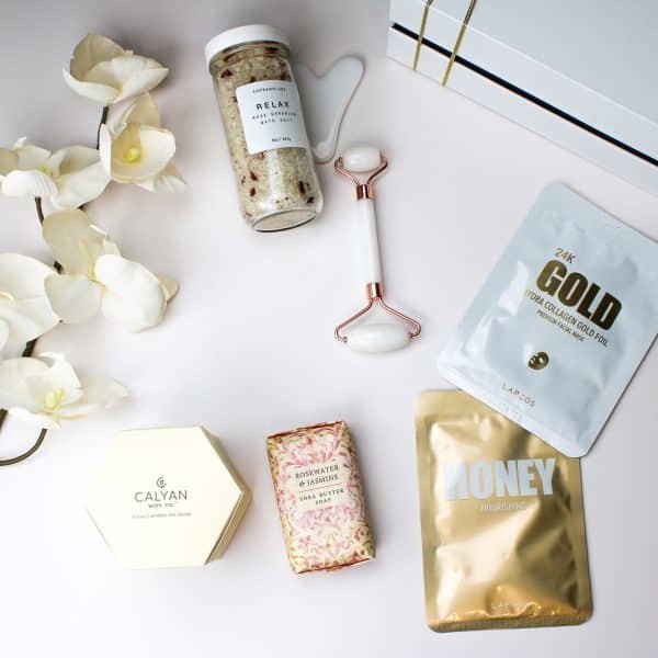 Flat lay of self-care essentials including rose geranium bath salt, jade roller, 24k gold and honey facial masks, Calyan candle, floral shea butter soap, and white orchid flowers